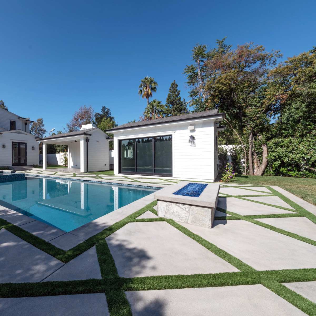 A modern backyard with a rectangular swimming pool surrounded by concrete and grass pathways, flanked by a pavilion and a covered seating area.