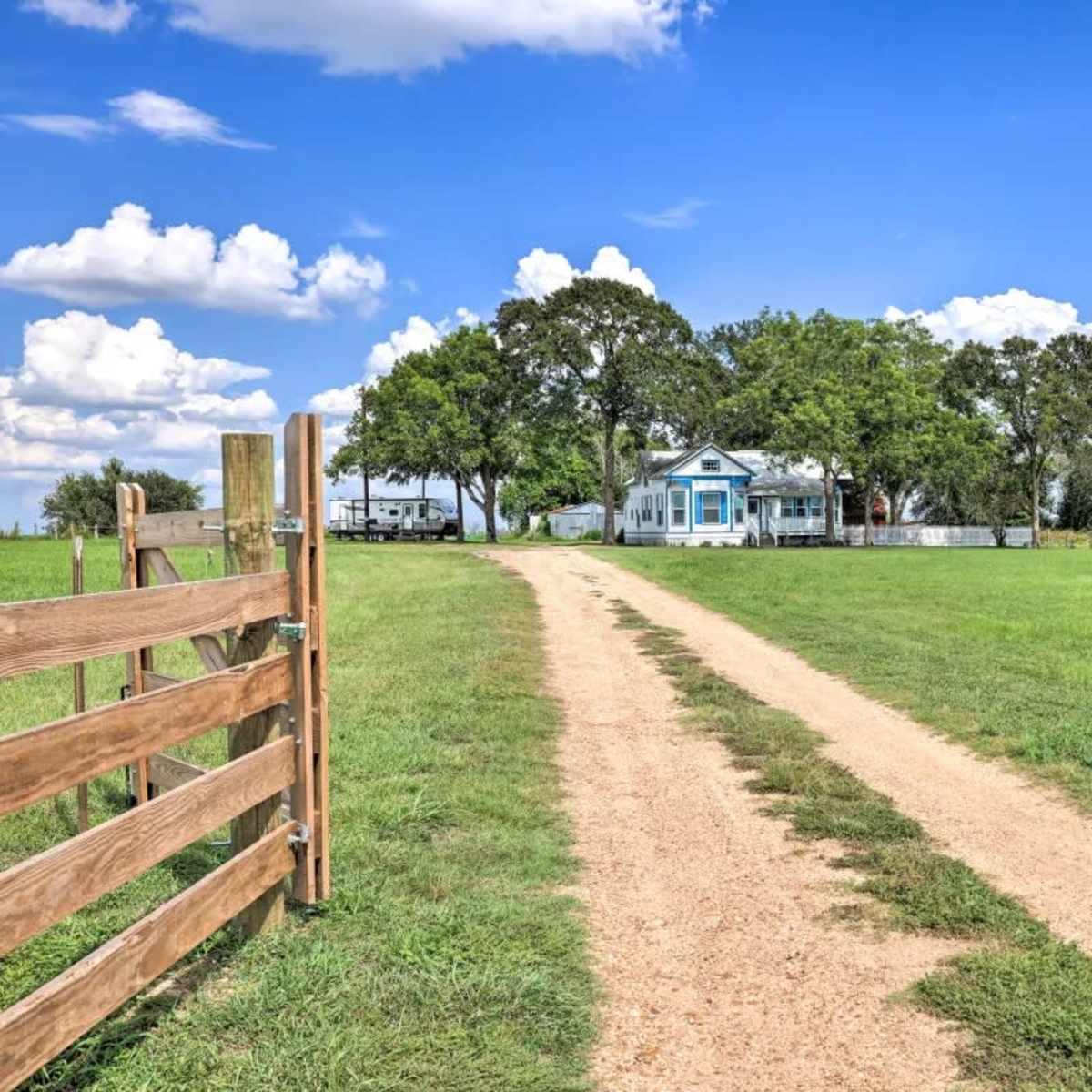A dirt driveway leads from a wooden gate towards a house surrounded by trees and open grassland under a blue sky with clouds.