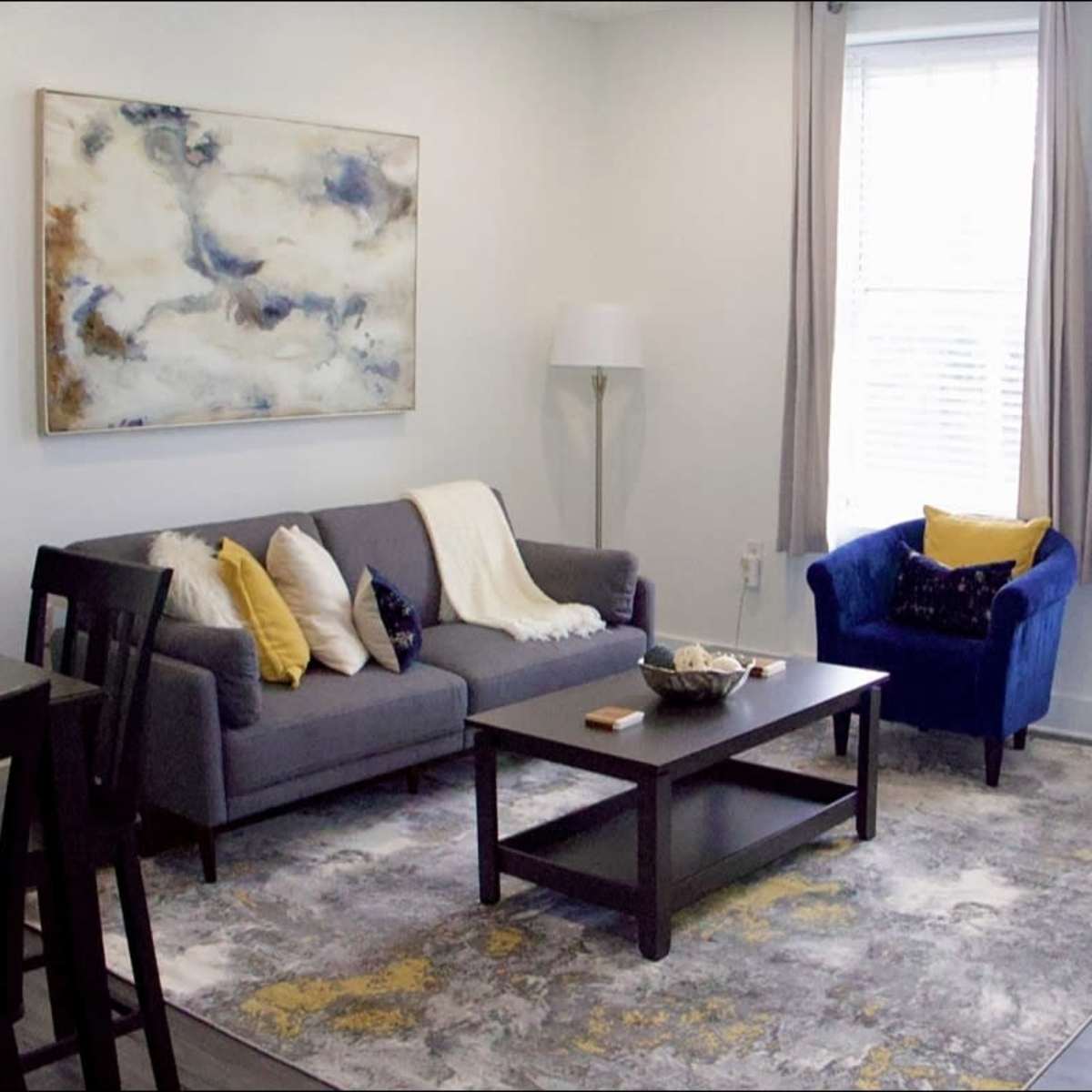 A modern living room features a gray sofa with decorative pillows, a blue armchair, a coffee table, and a large abstract painting on the wall, illuminated by natural light from two windows.