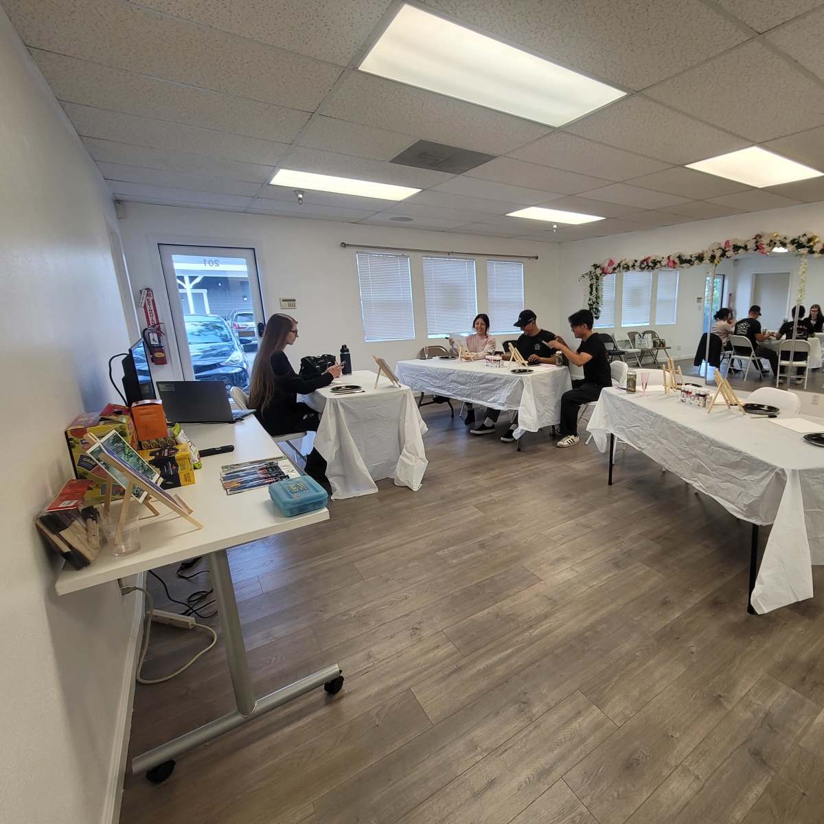 The room features several tables covered with white tablecloths, where individuals are engaged in activities, and a floral arrangement decorates the wall.