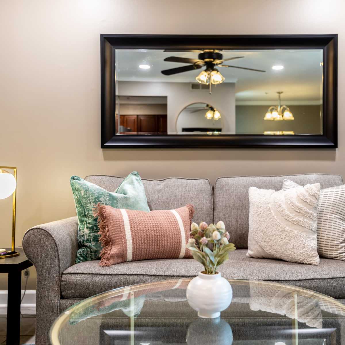 A gray sofa with various decorative pillows sits in front of a large mirror, reflecting light fixtures and a kitchen area in a living room.