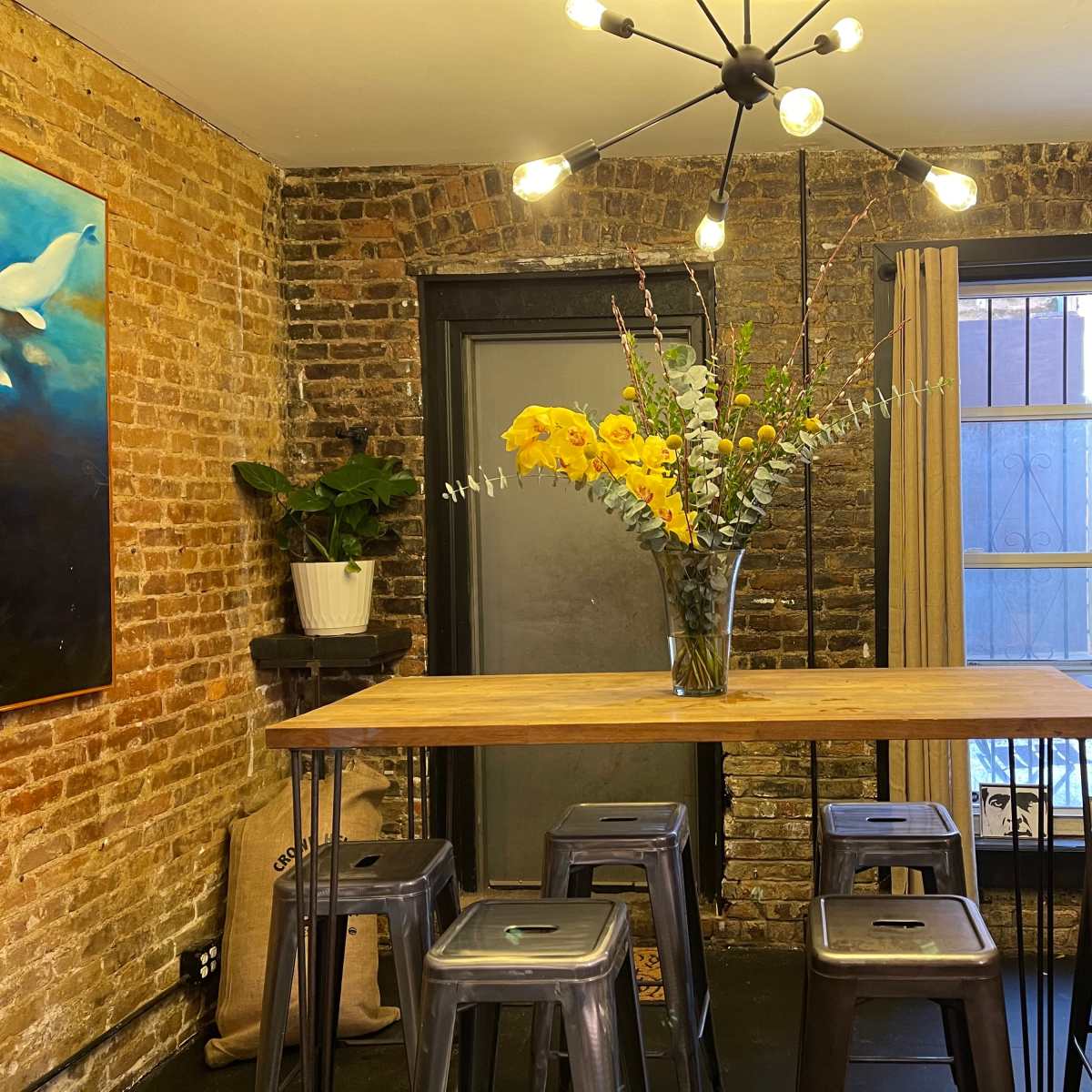 A modern dining space features a wooden table with metal stools, surrounded by exposed brick walls, potted plants, and a vibrant floral centerpiece under a contemporary light fixture.