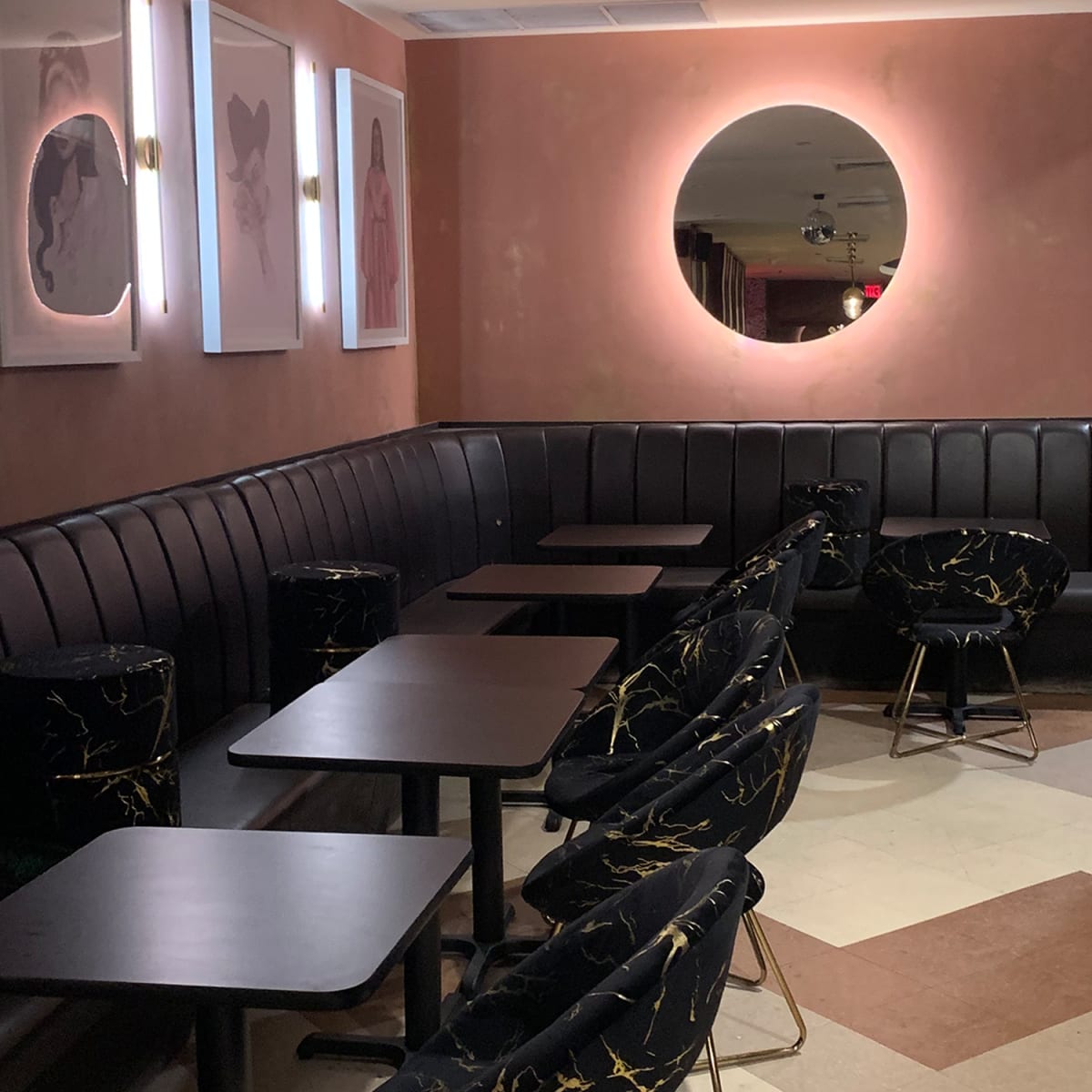 A dimly lit interior of a café or lounge featuring black seating with gold accents, round tables, and a circular mirror on the wall.
