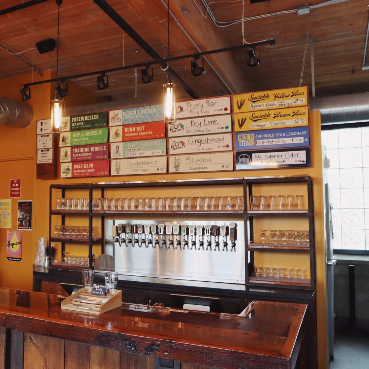 A bar area with a wooden counter, a wall featuring a menu of beverages, and multiple beer taps.