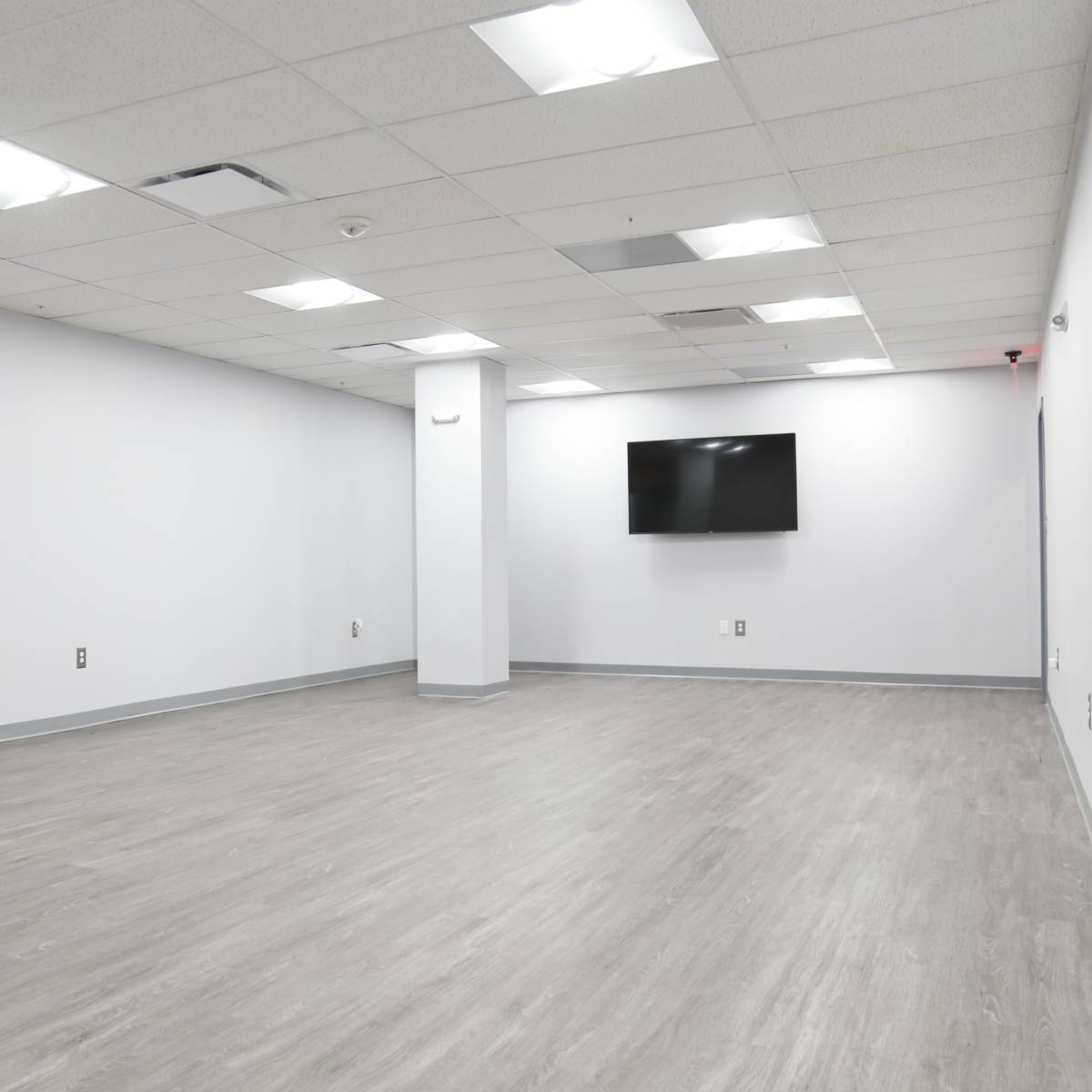 An empty, well-lit room with gray flooring, white walls, a wall-mounted television, and no furniture.
