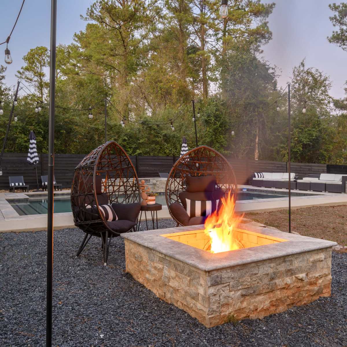 A backyard area with two hanging chairs near a stone fire pit, surrounded by landscaping and a swimming pool in the background.