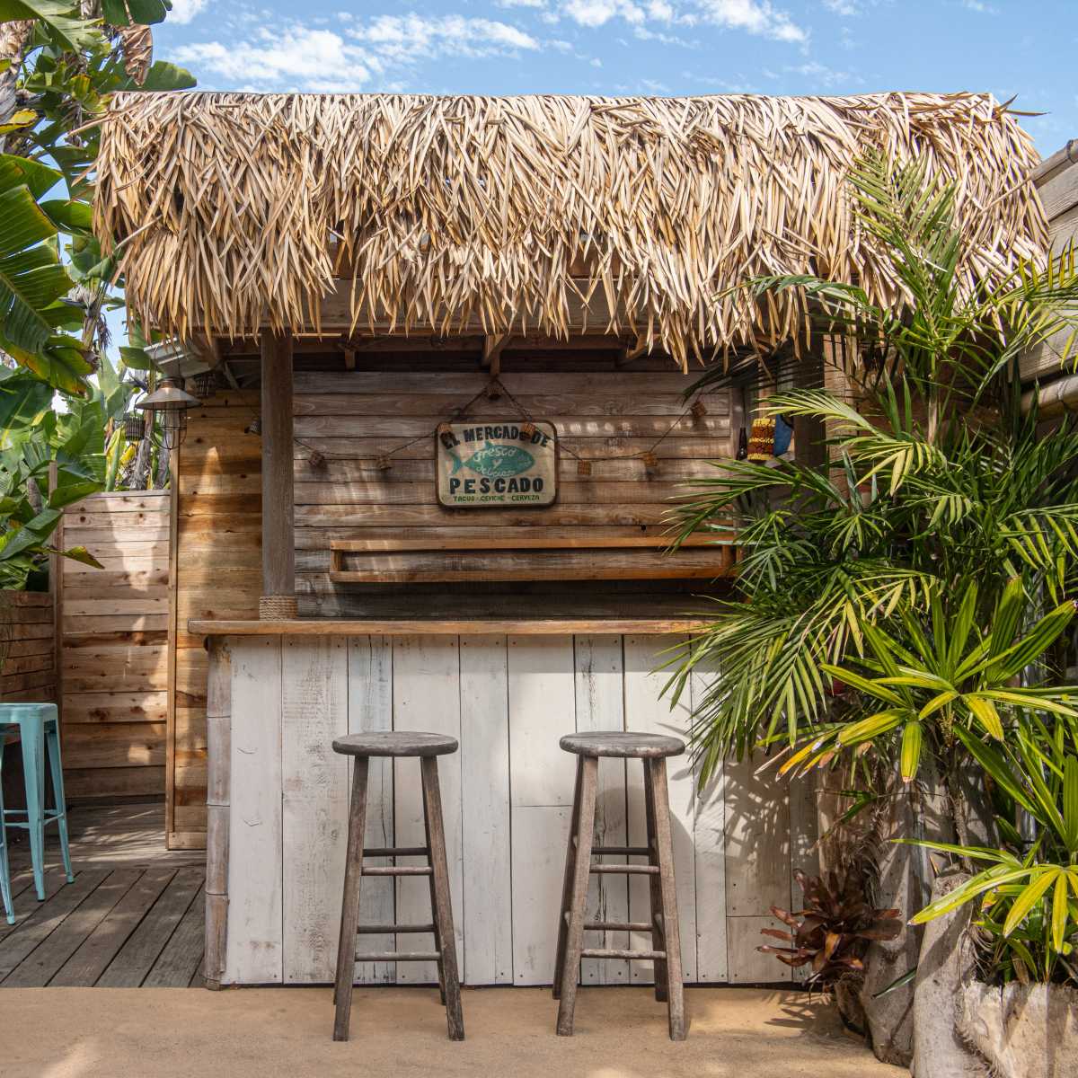 A rustic outdoor bar with a thatched roof, wooden counter, and two tall stools surrounded by tropical plants.
