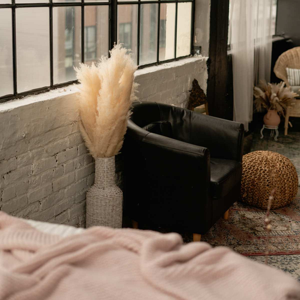 A cozy interior space featuring a black chair, a large beige pampas grass arrangement in a woven vase, and large windows with urban views.