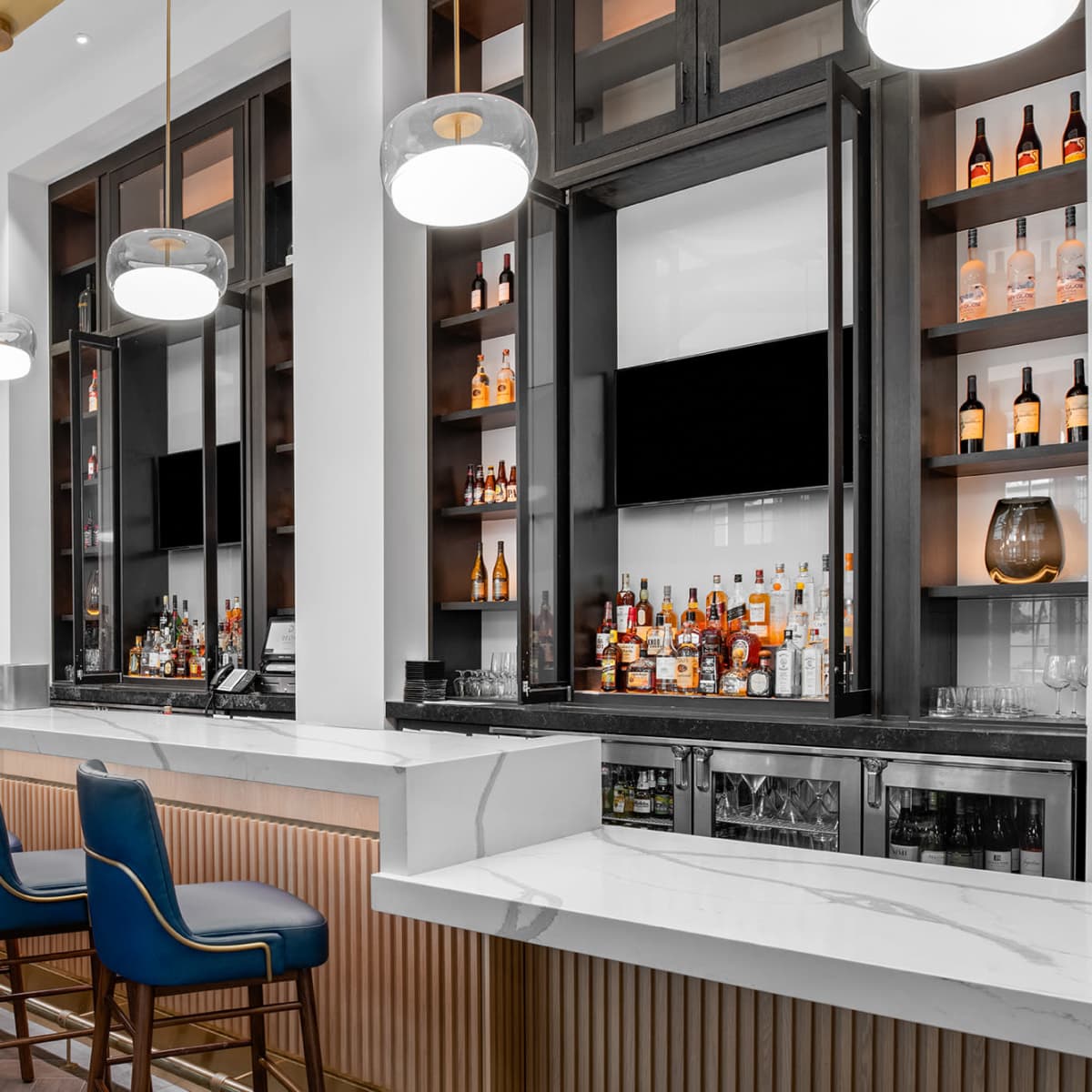 A modern bar area featuring a long marble counter, high-backed blue chairs, and shelves with various bottles and decor.