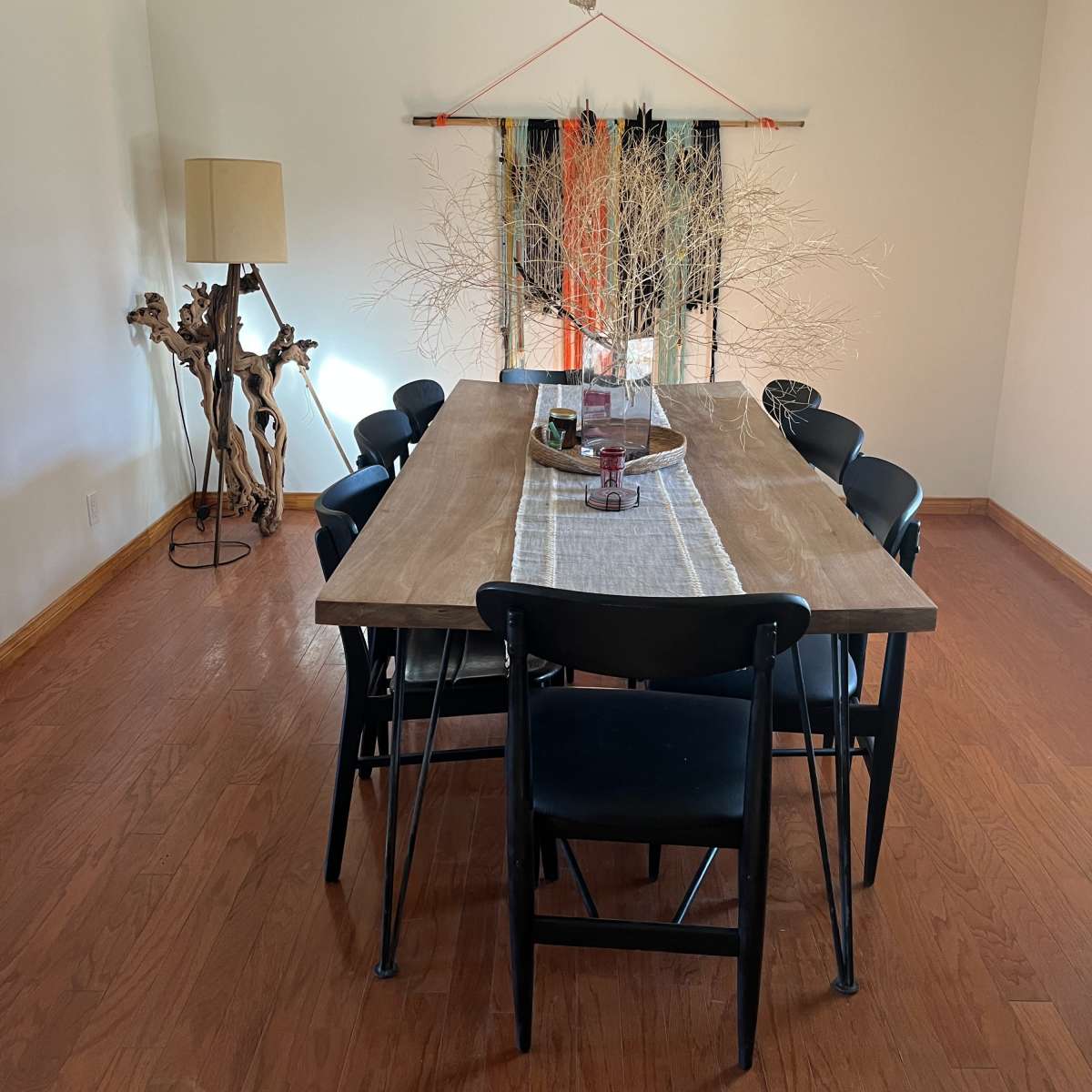 A wooden dining table surrounded by black chairs is set in a bright room with a floor lamp and decorative branches in the center.