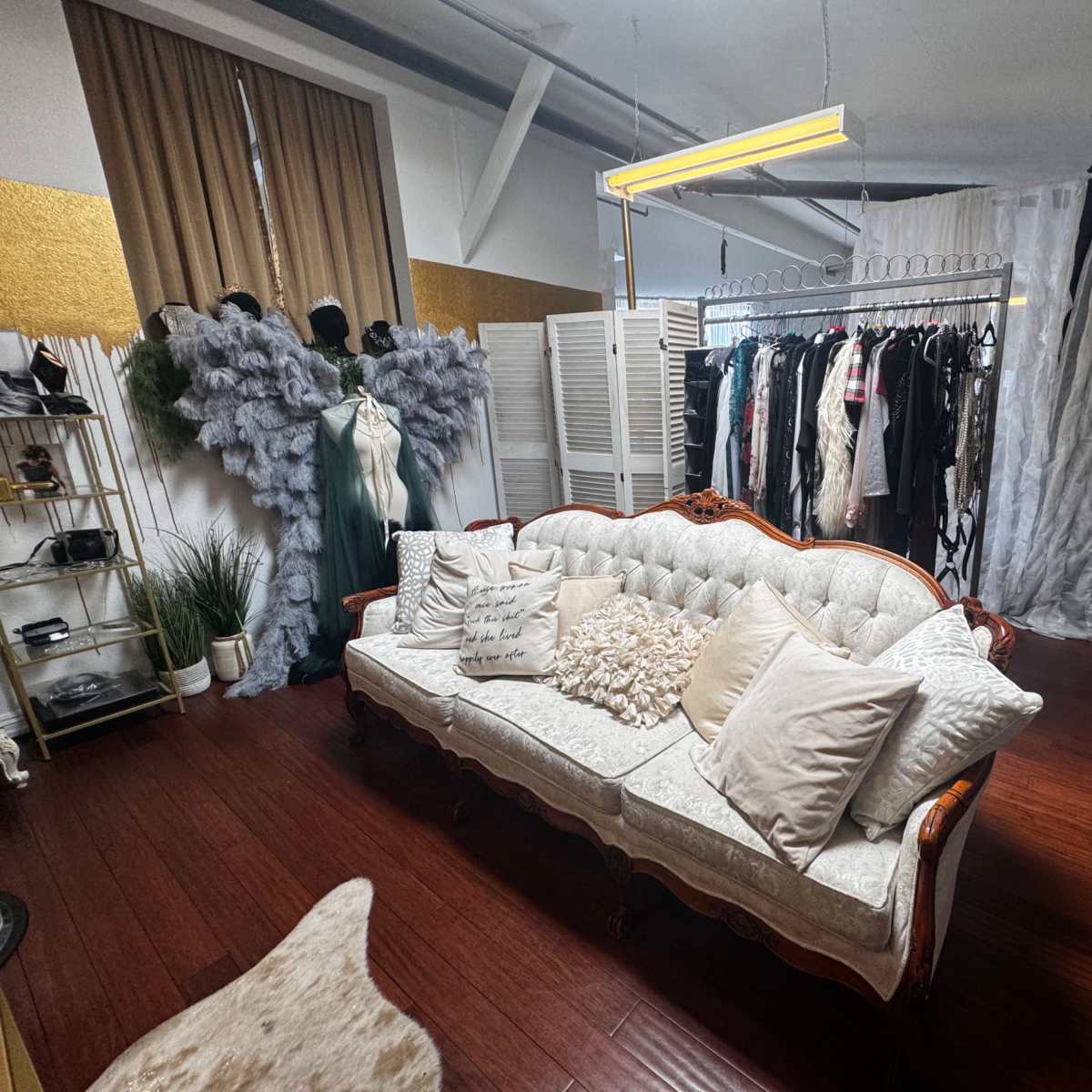 A styled studio space featuring a vintage sofa with decorative pillows, clothing racks filled with various garments, and draped fabric backgrounds.
