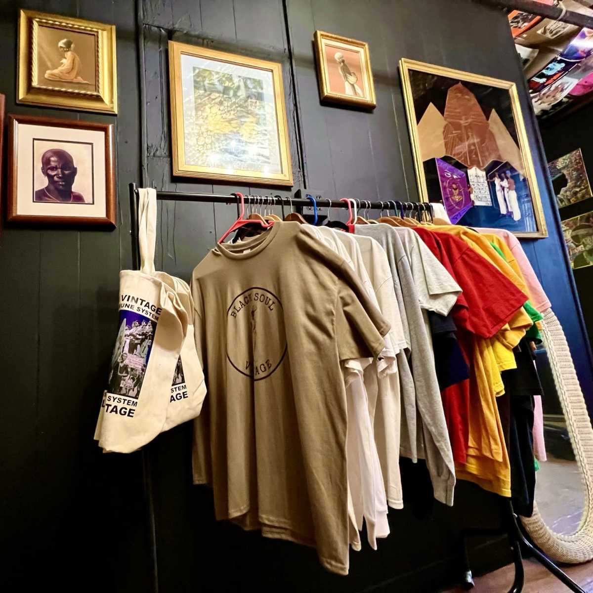 A rack of assorted t-shirts and a tote bag hangs against a dark wall adorned with various framed artworks.