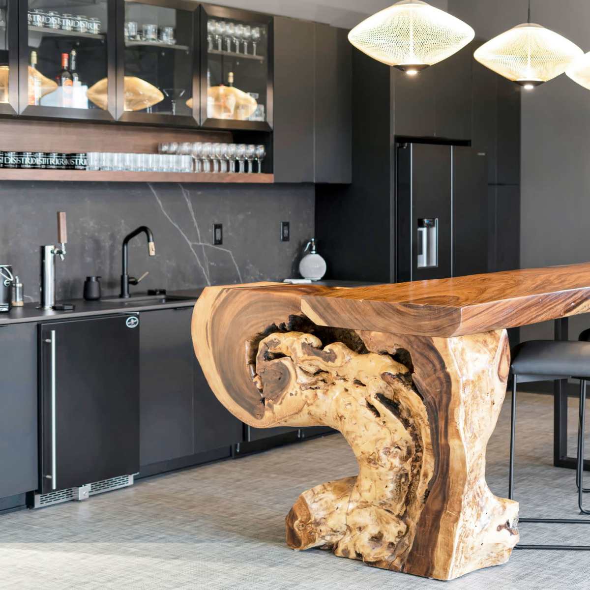 A modern kitchen bar featuring a large wooden countertop supported by a sculptural base, with four black stools positioned along one side.