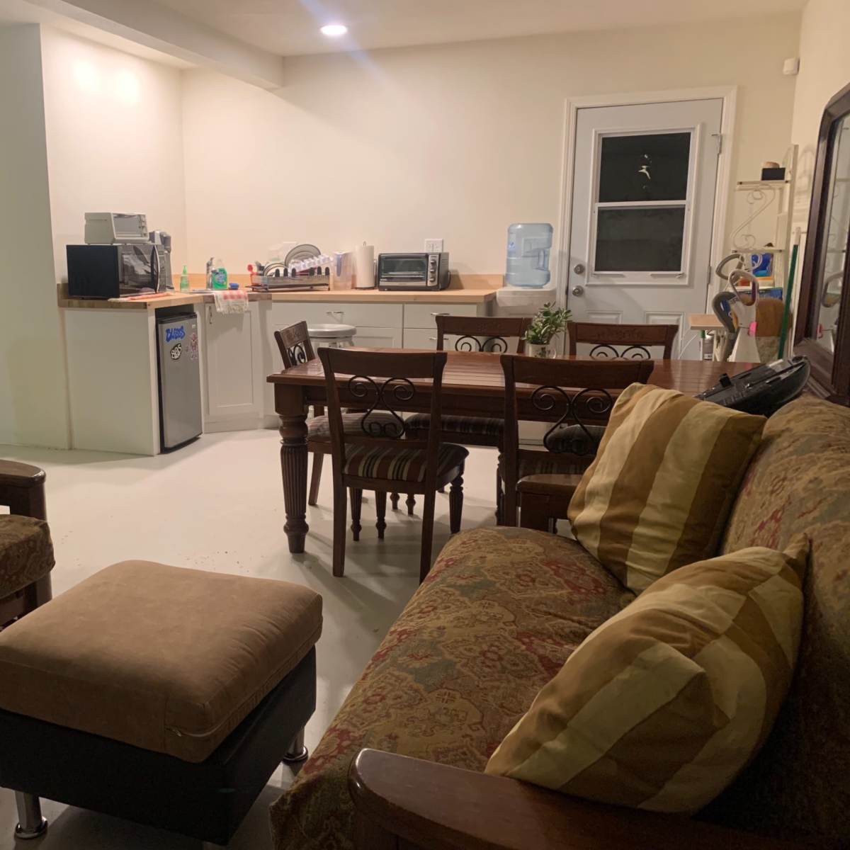 A living area with two couches, an ottoman, and a dining table near a kitchenette with a refrigerator and various appliances.