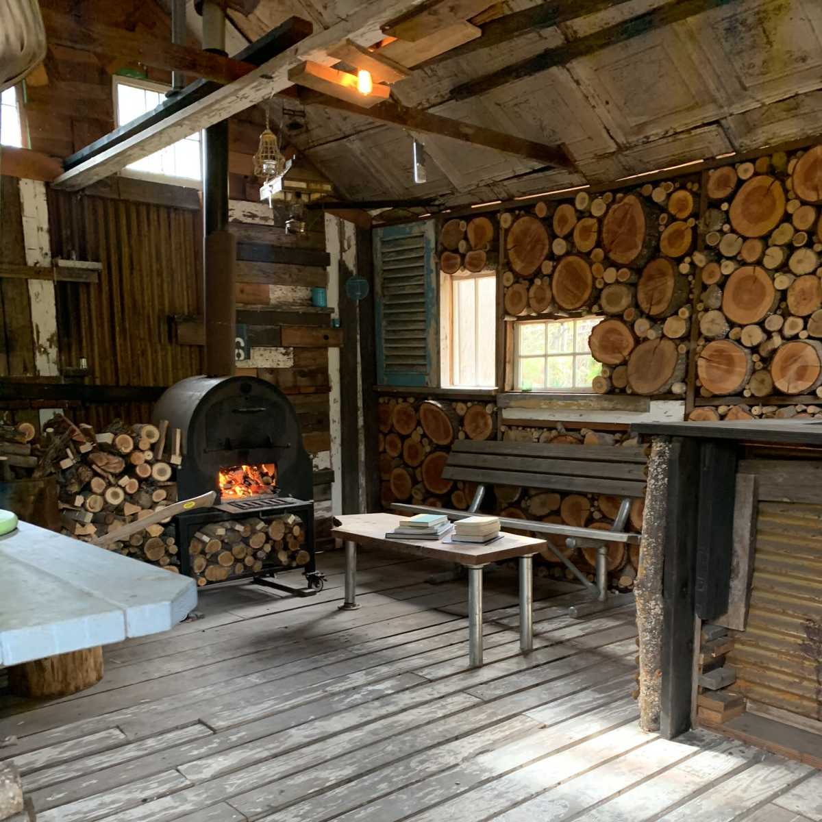 The interior of a rustic cabin features stacked firewood, a stove with a fire, and a simple wooden table.