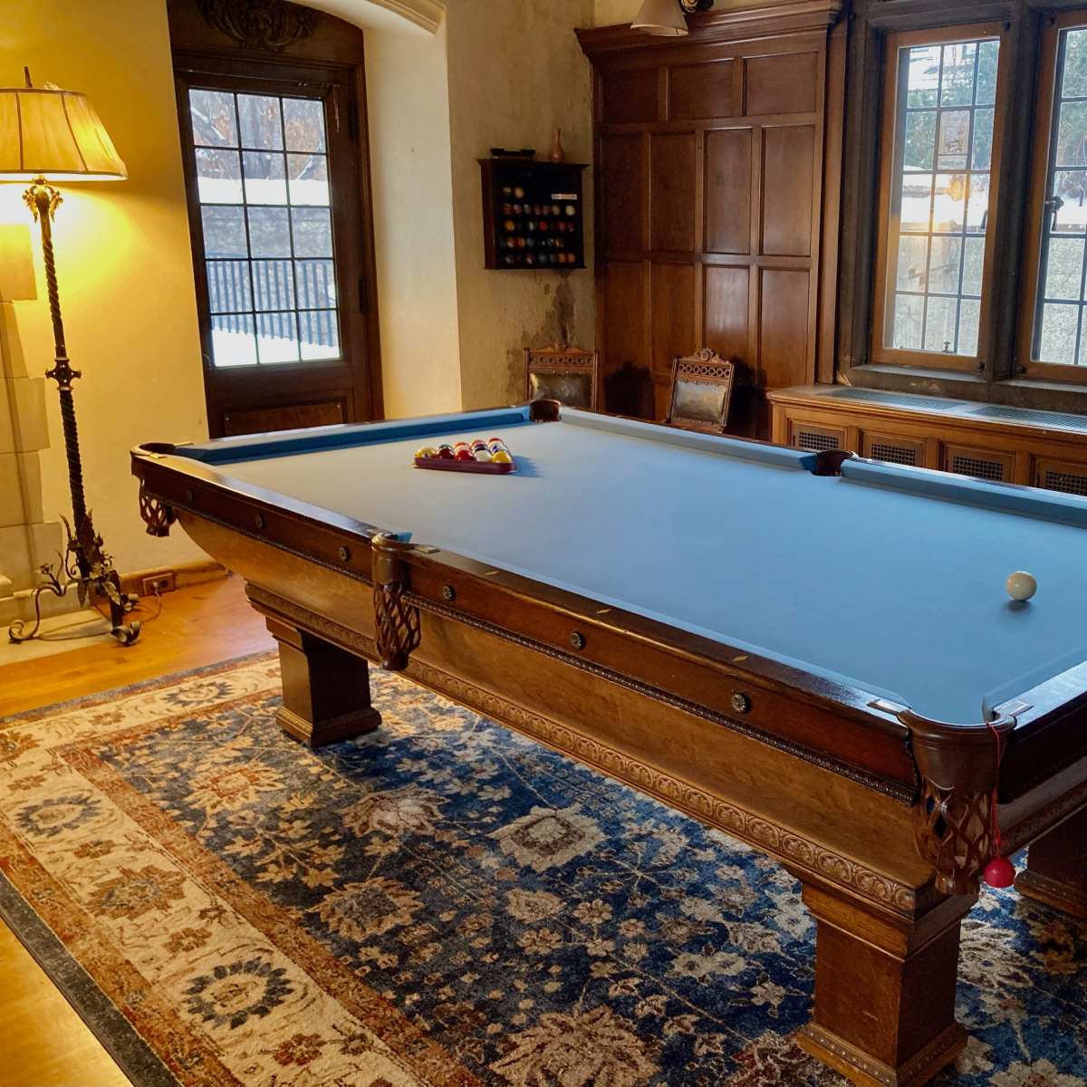 A wooden billiard table with a blue felt surface is positioned in a room featuring large windows and warm wood paneling.