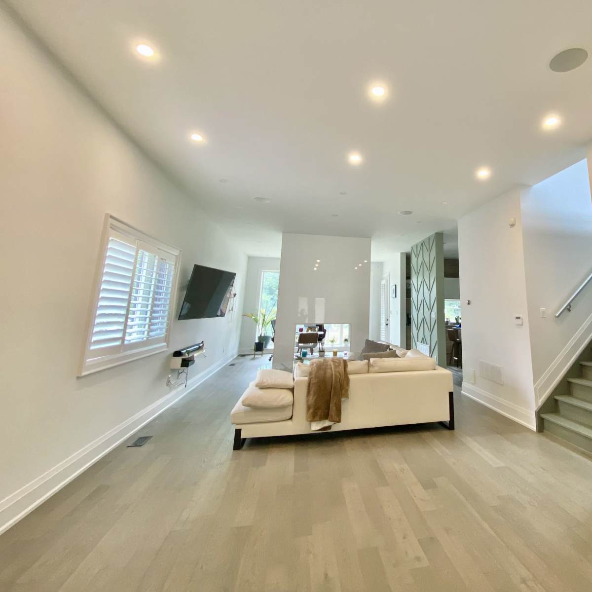 A modern living room features a white sofa, a wall-mounted television, and a piano, with stairs leading to an upper level.
