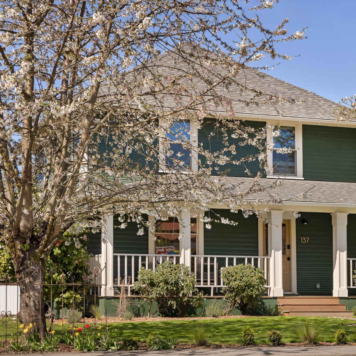 A two-story green house with a front porch is surrounded by blooming trees and a decorative fountain in the yard.