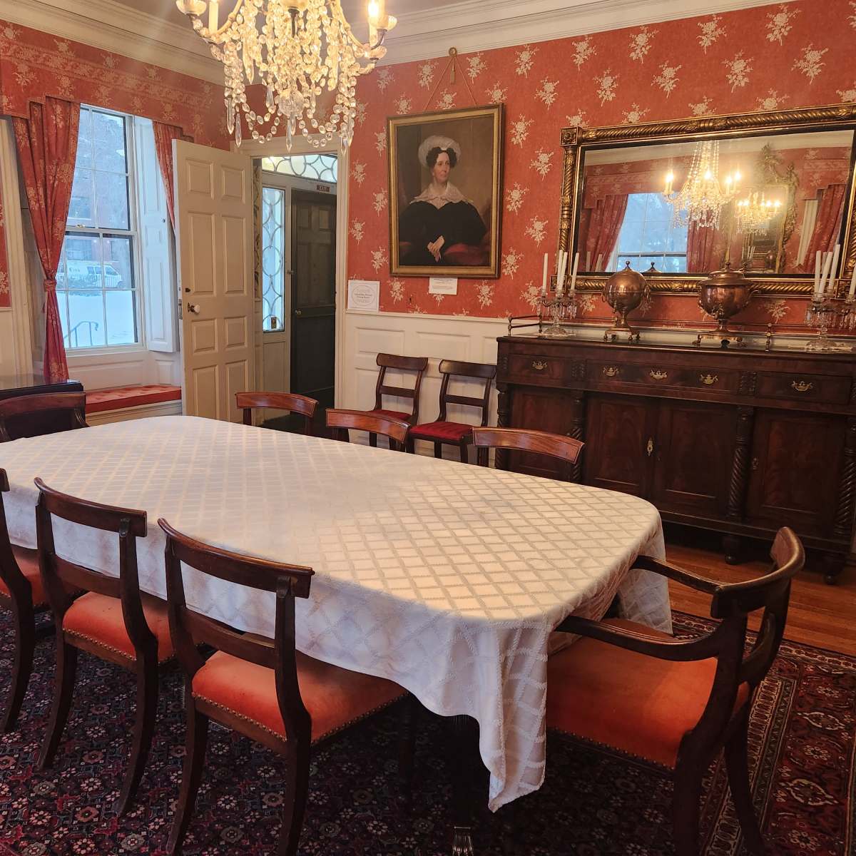 A dining room with a large table surrounded by chairs, ornate wallpaper, and a chandelier, featuring a mirror and decorative items on a sideboard.