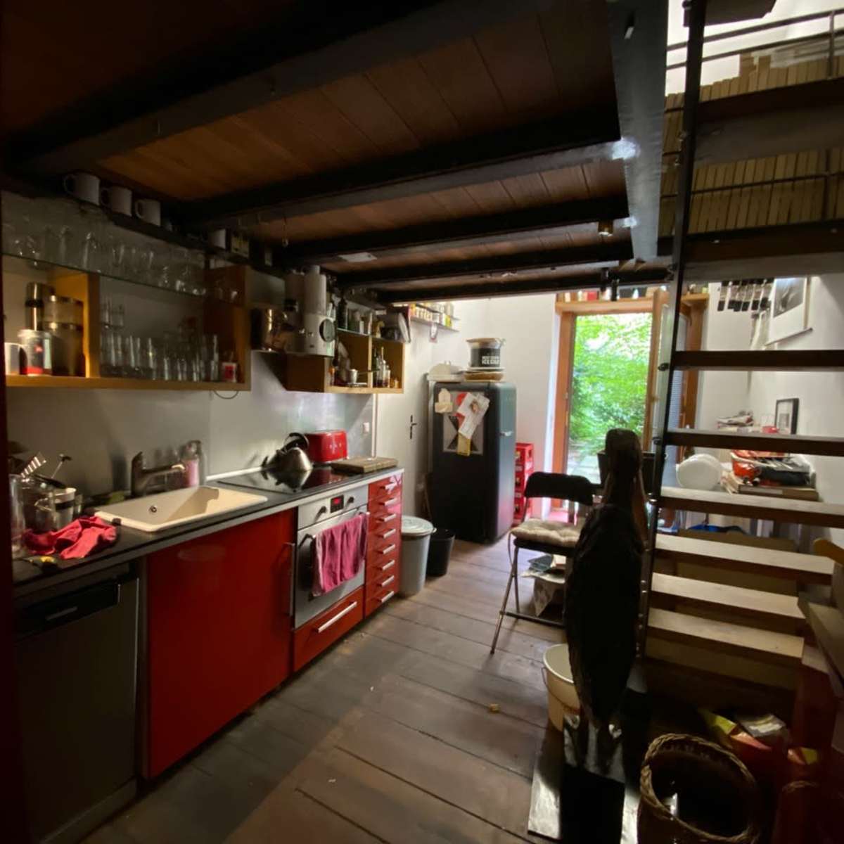 A compact kitchen with red cabinets, stainless steel appliances, and wooden beams, leading to a stairway and an open doorway filled with greenery.