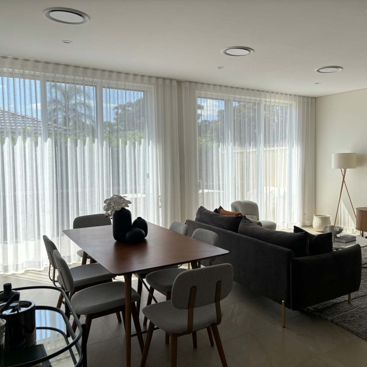 A modern living room with large windows covered by sheer curtains, a dining table, a sofa, and a television on a low cabinet.