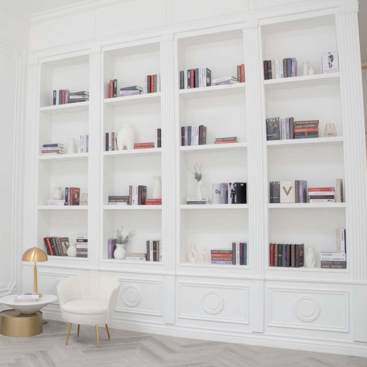 A modern interior space featuring a white bookshelf filled with books, a small round table, a chair, and a desk with a lamp against a light-colored wall.