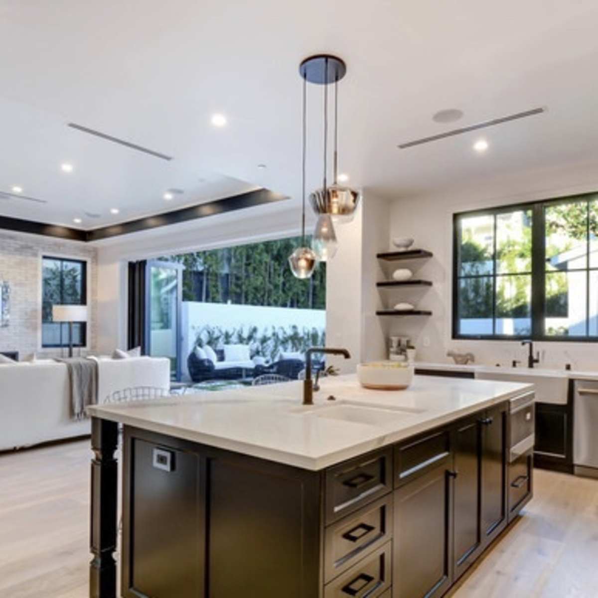 A modern kitchen with an island, stainless steel appliances, and a view of an outdoor seating area through large windows.