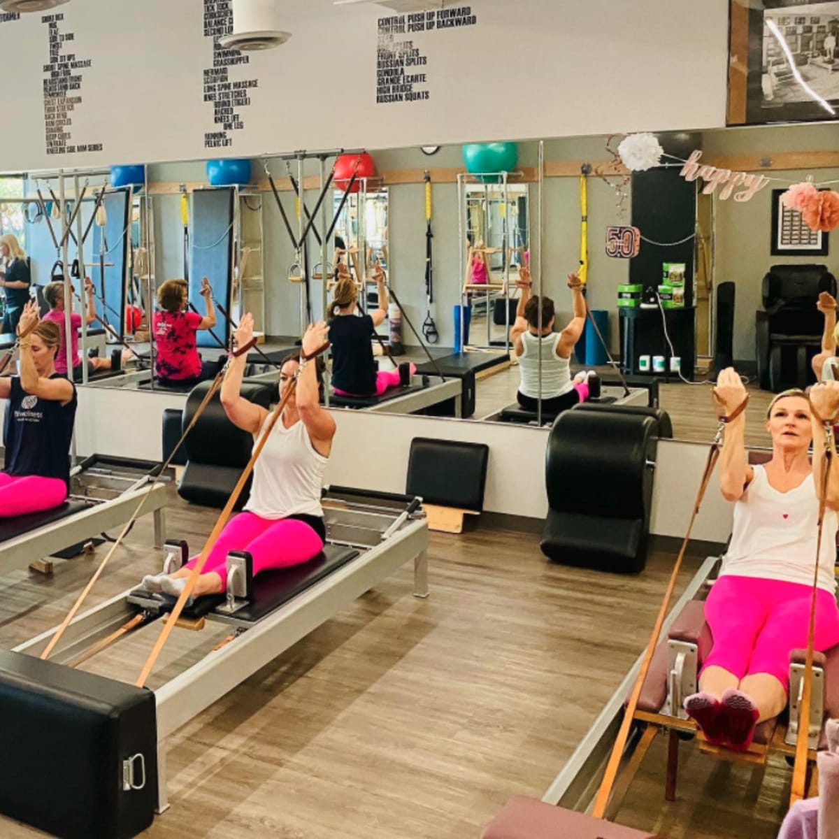 A group of women participate in a rowing exercise class in a fitness studio with mirrors and gym equipment.