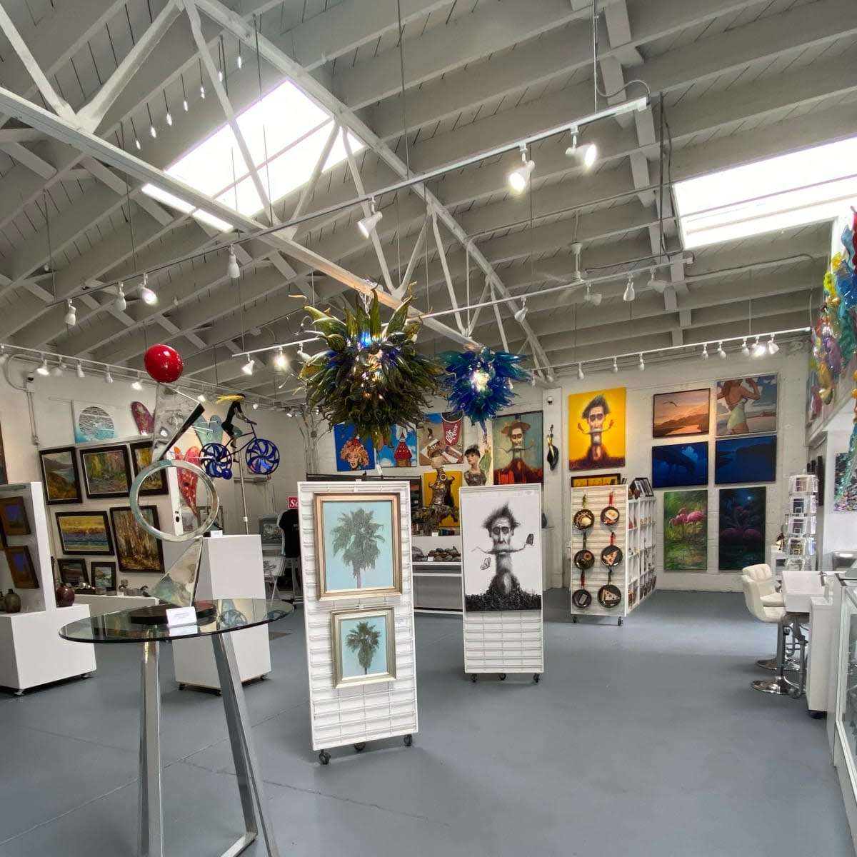 The interior of an art gallery featuring various artworks displayed on white walls, with sculptures and decorative pieces arranged throughout the space.