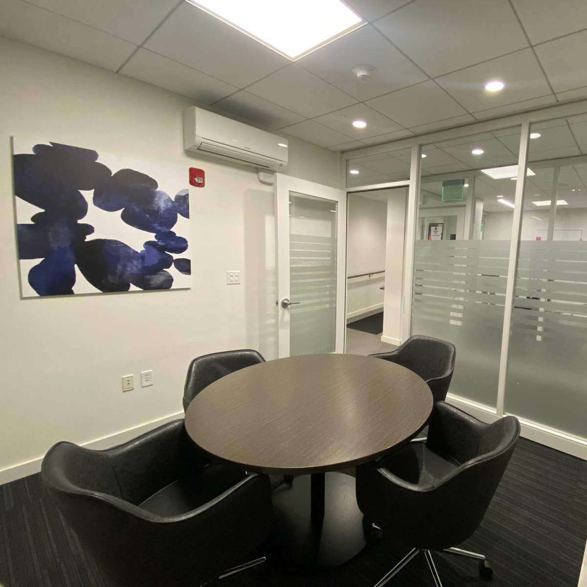 A modern office meeting room with a round table and four chairs, separated by glass walls from the adjacent area.