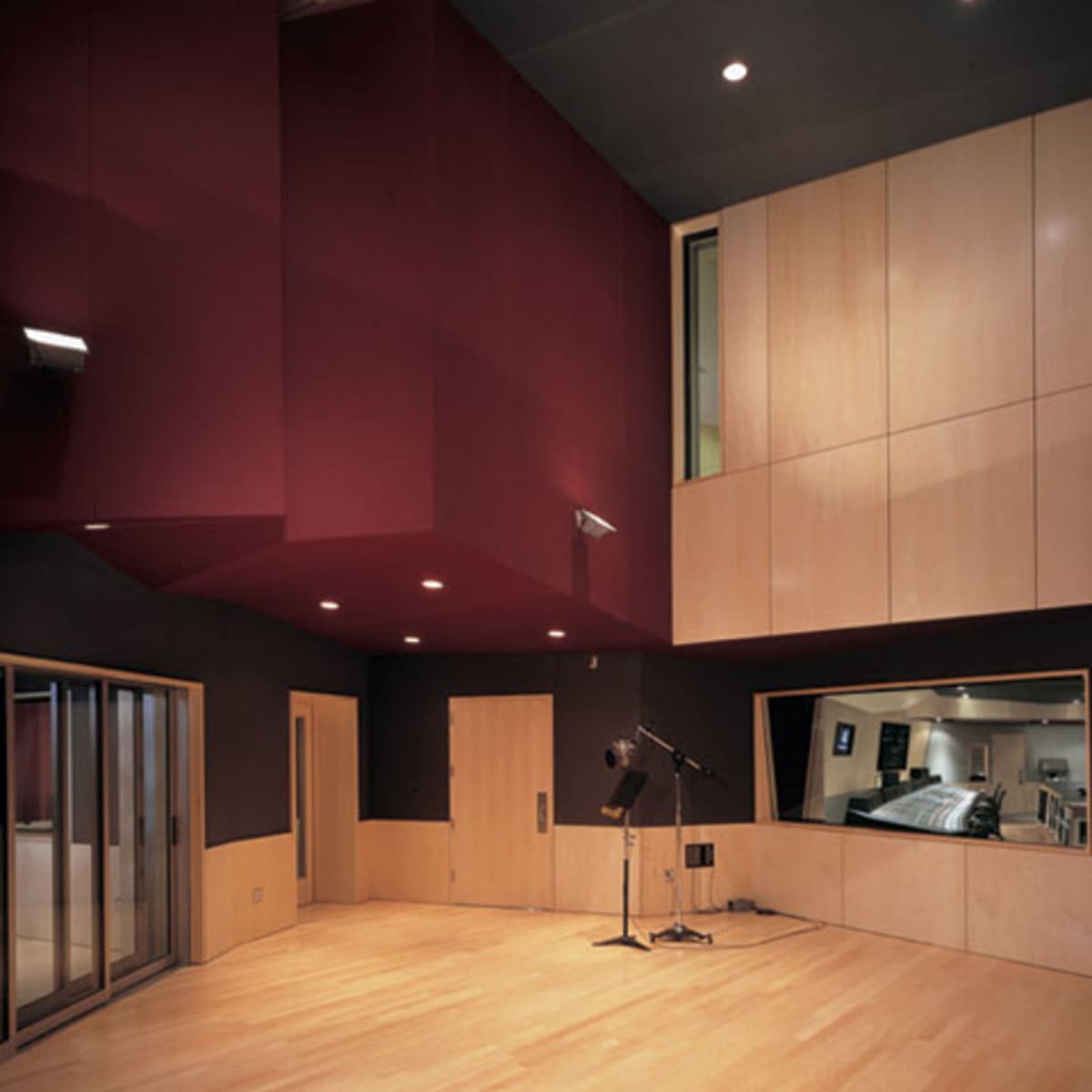 A modern recording studio with a wooden floor, red and black walls, and a large window overlooking another room.