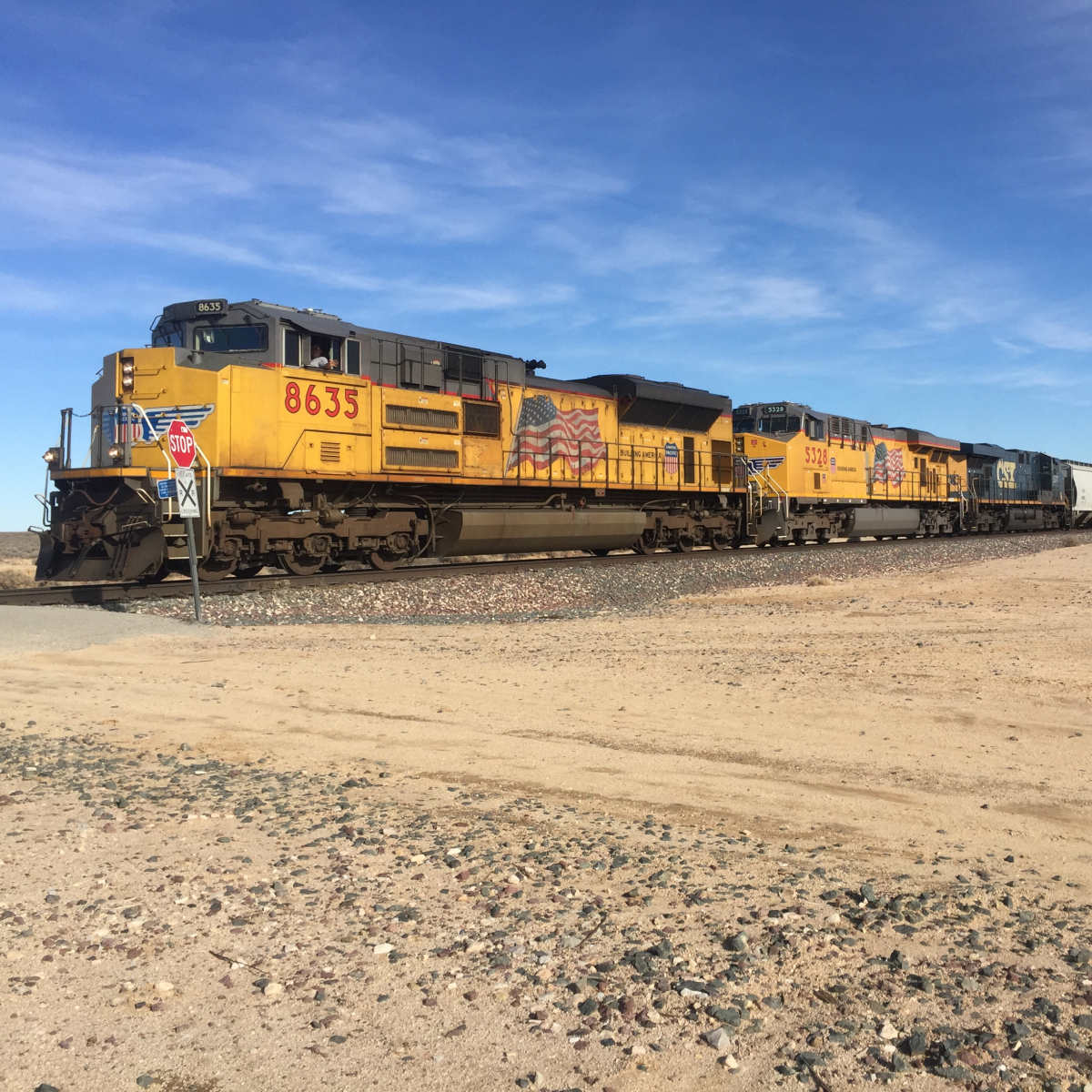 A freight train with yellow locomotives is passing through a desert landscape, with a clear blue sky overhead.