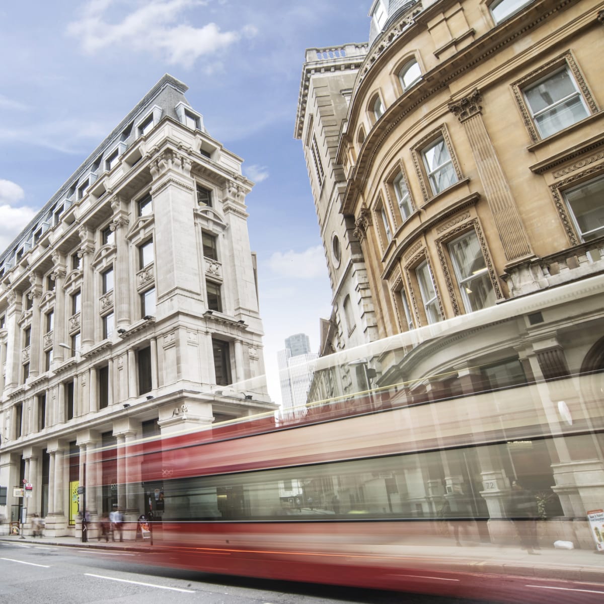 A city street with historic buildings on either side and a blurred red bus moving through the scene.