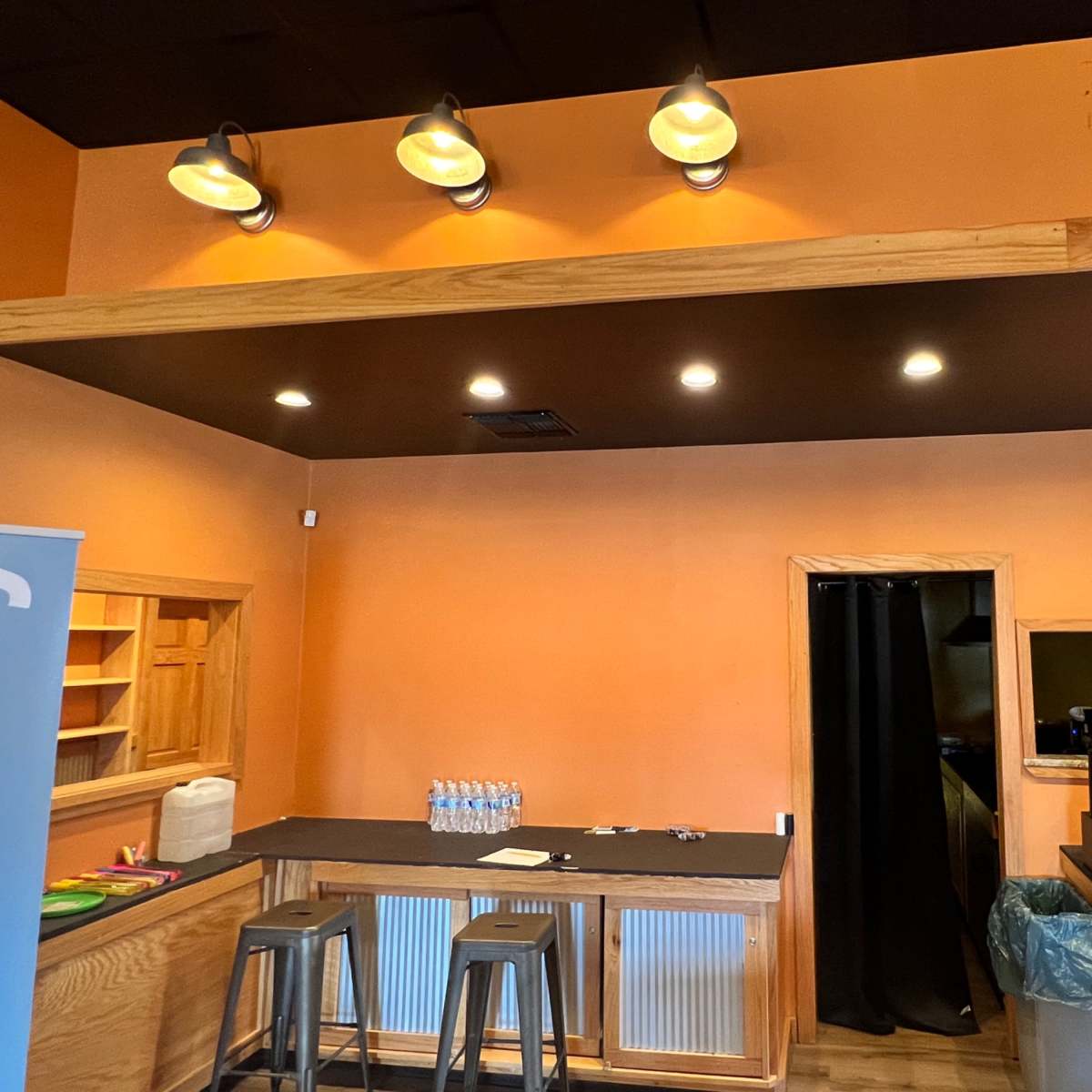 A bright orange-walled café interior with a wooden bar counter, bar stools, and a small kitchenette area.
