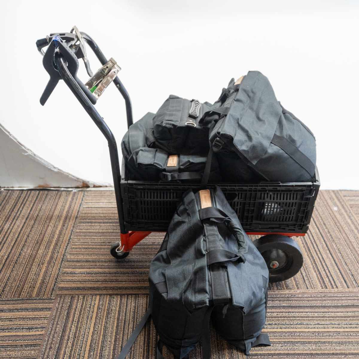 A black cart holds several large black backpacks on a striped carpet surface.