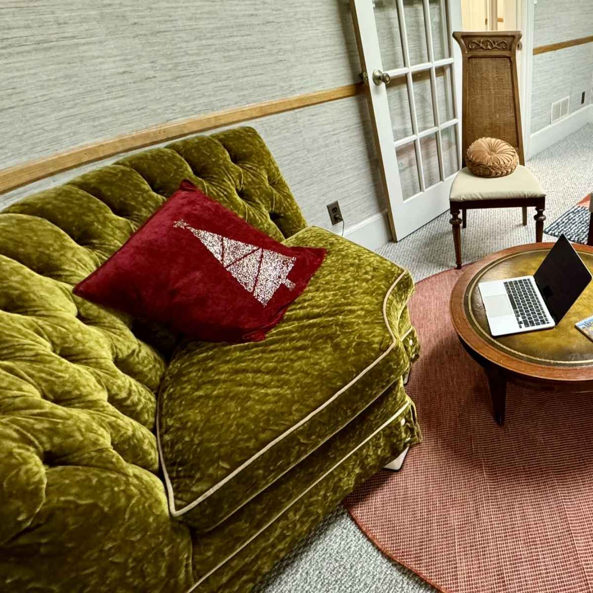 A cozy sitting area features a green velvet sofa, a round coffee table with a laptop, and several upholstered chairs arranged on a patterned rug.