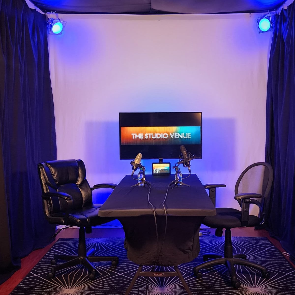 A recording studio setup with two chairs flanking a table, a backdrop of black curtains, and a screen displaying the text "THE STUDIO VENUE."