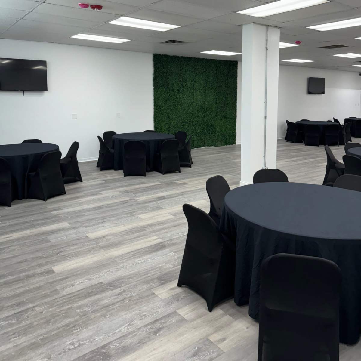 A spacious room with several round black tables covered with black tablecloths and a green wall of plants at one end.