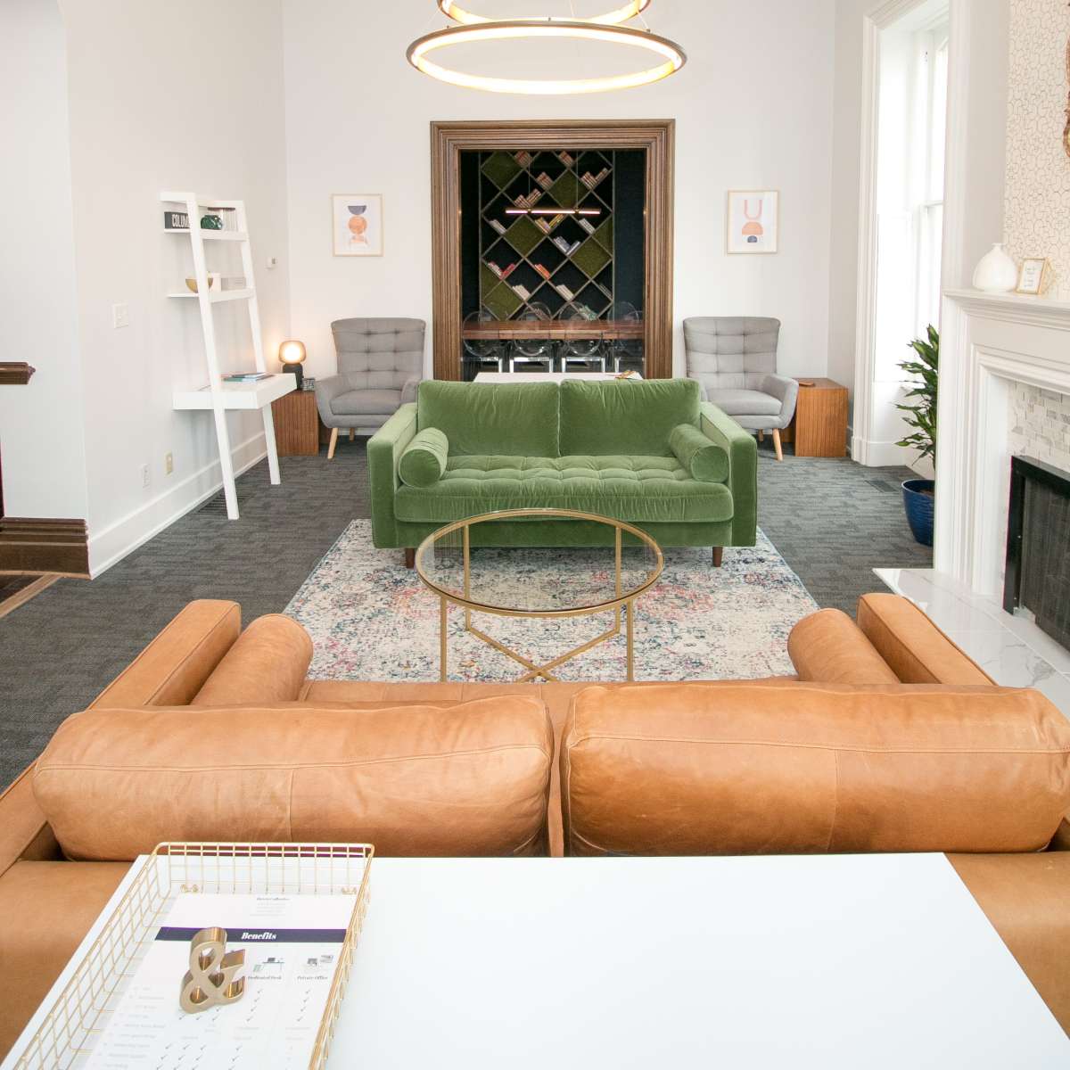 A modern living room with a green velvet sofa, two gray armchairs, a circular chandelier, and a wine rack in the background.