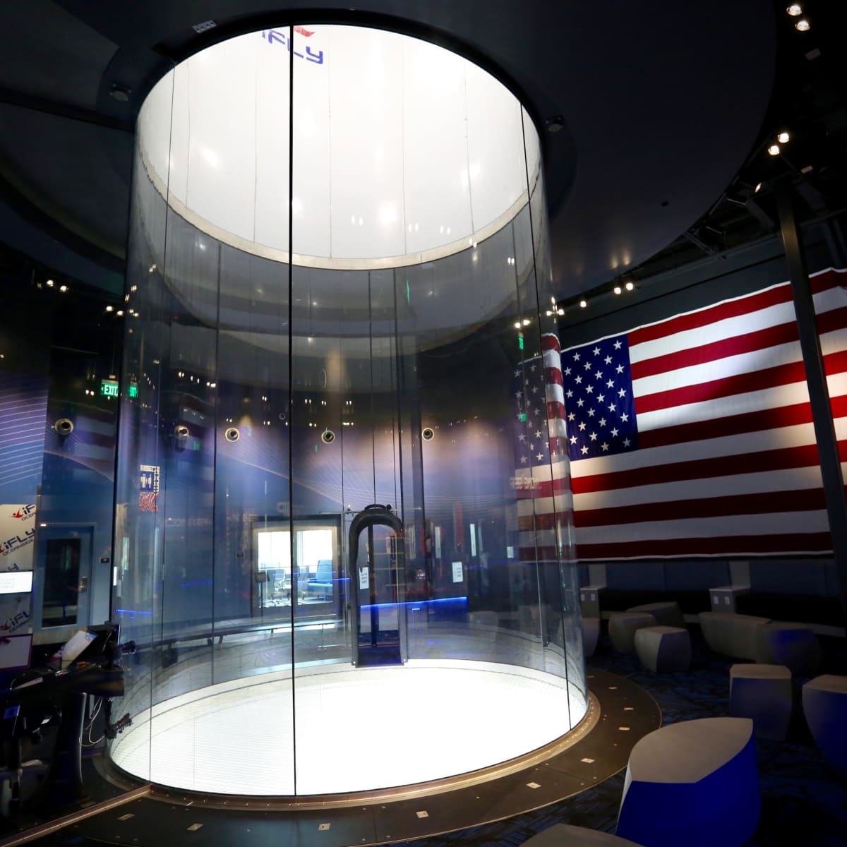 A spacious indoor skydiving facility featuring a large transparent flight chamber and a prominent American flag on the wall.