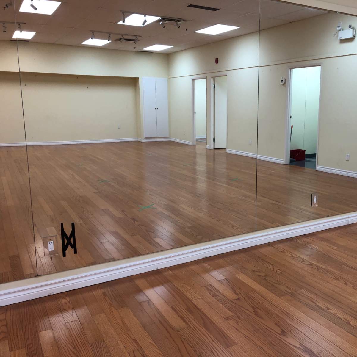 An empty room with wooden flooring, a large mirror along one wall, and a white door on the opposite side.