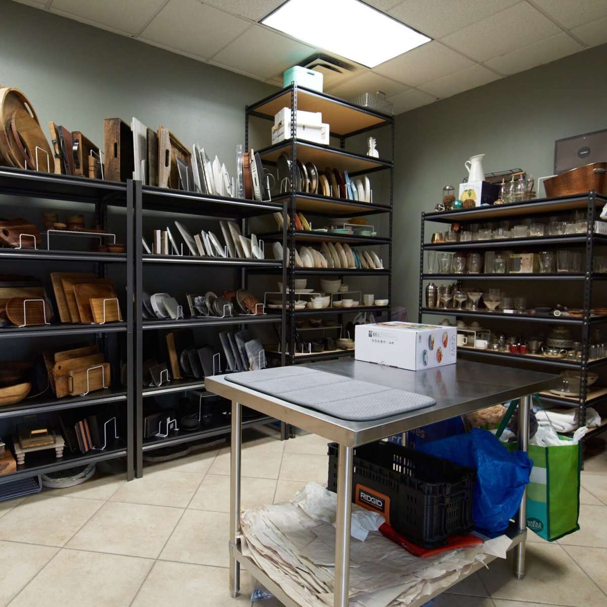 A well-organized storage room features metal shelves filled with wooden and ceramic kitchenware, alongside a table in the center.
