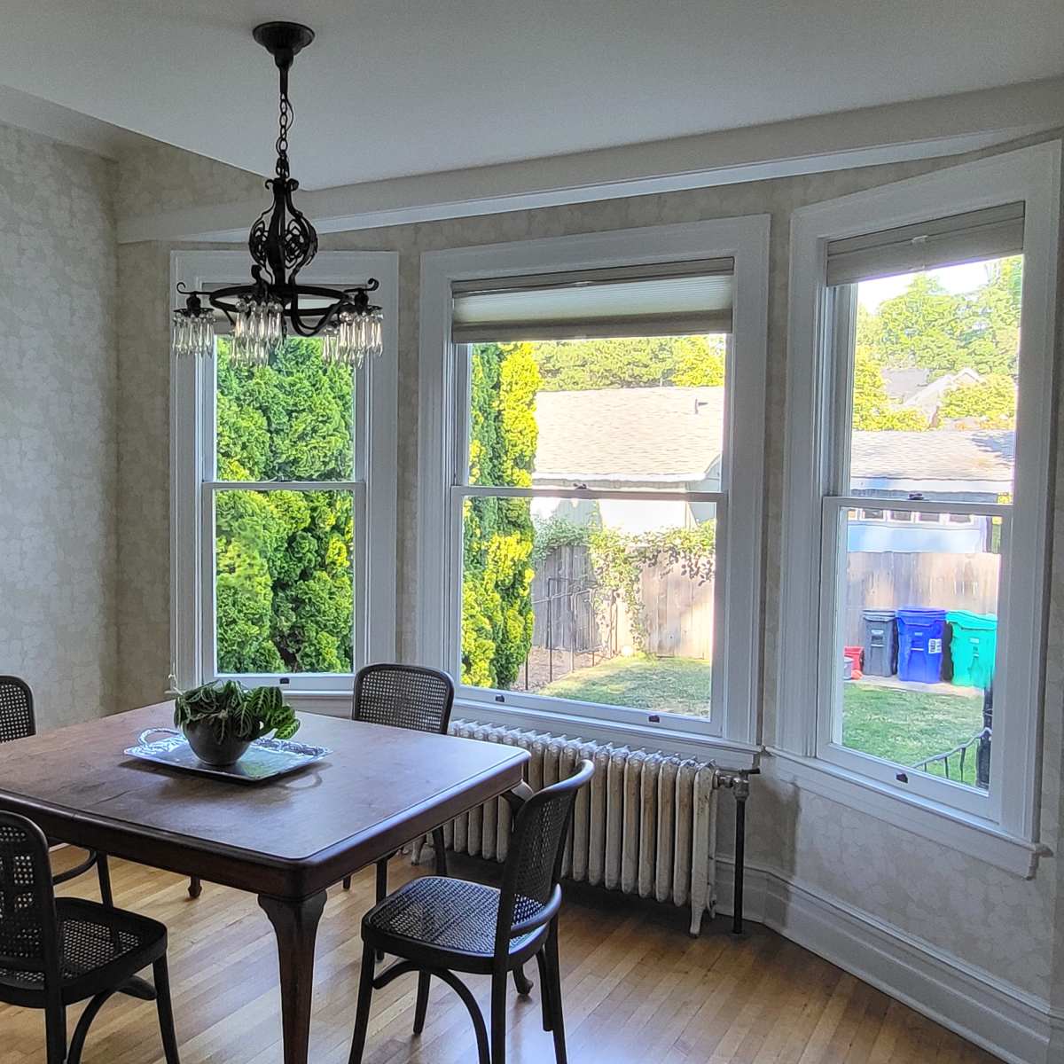 A dining area features a wooden table with four chairs, illuminated by a chandelier, with large windows showing green foliage outside.