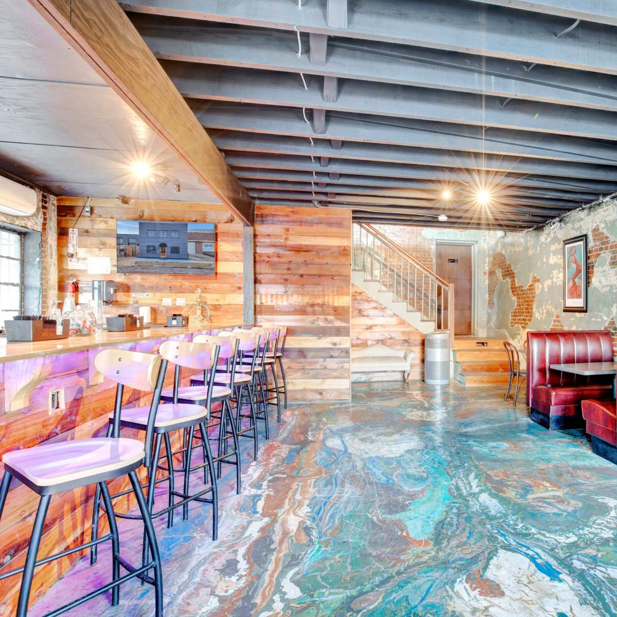 A contemporary bar area with wooden walls, marbled flooring, and red leather booths, leading to a staircase in the background.