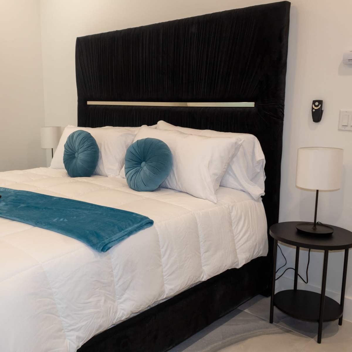 A neatly made bed with a black upholstered headboard, blue accent pillows, and a light blue throw blanket, positioned near a nightstand with a lamp.