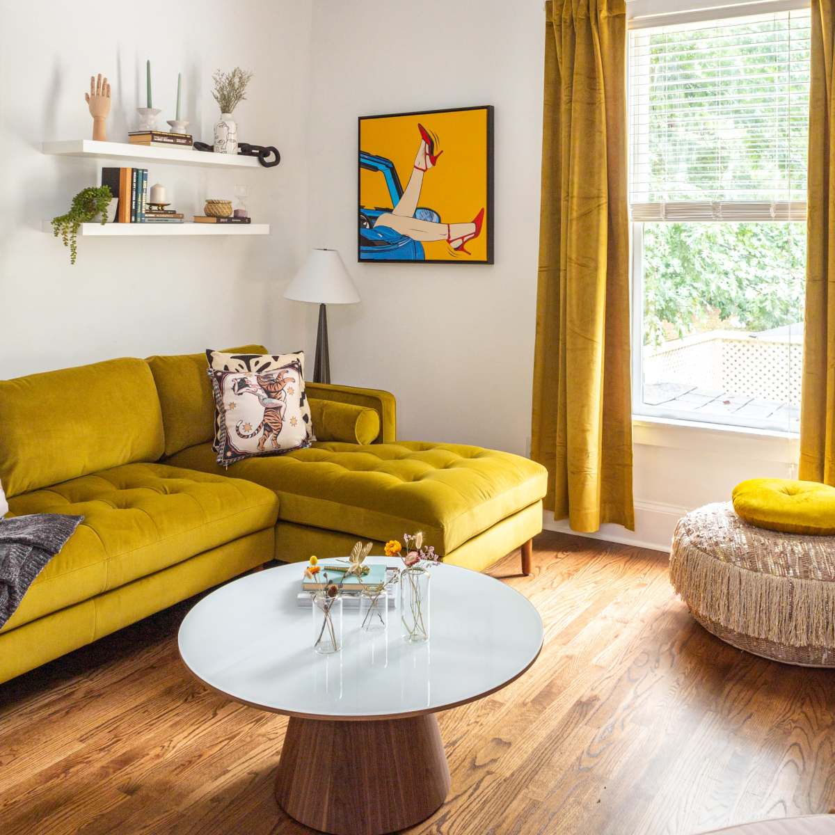 A modern living room featuring a yellow sofa, a round coffee table, a bright artwork on the wall, and large windows with yellow curtains.