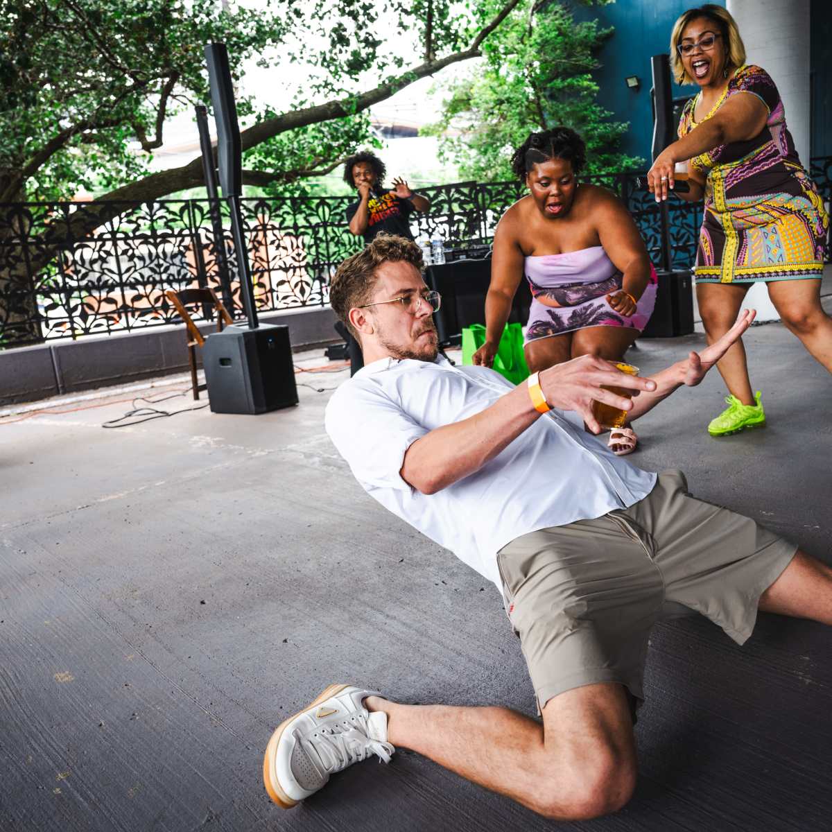 A man in a white shirt and shorts performs a dance move on the floor while two women cheer him on in a lively outdoor setting.