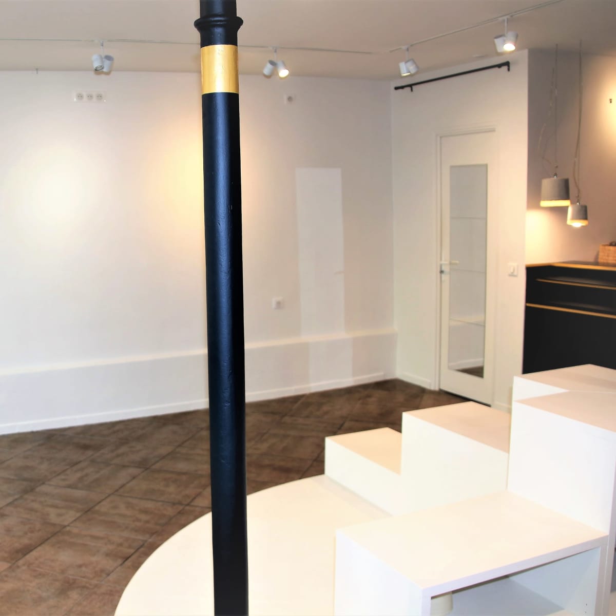 An empty room with white walls, a circular white platform, and a black support column in the center, illuminated by track lighting.