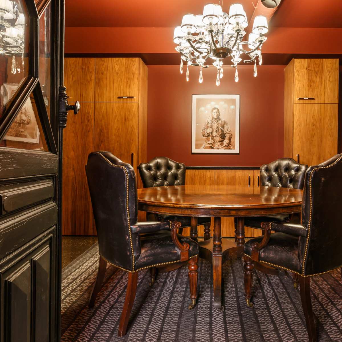 A wooden dining table with four black upholstered chairs is set in a dimly lit room adorned with dark red walls and a chandelier.