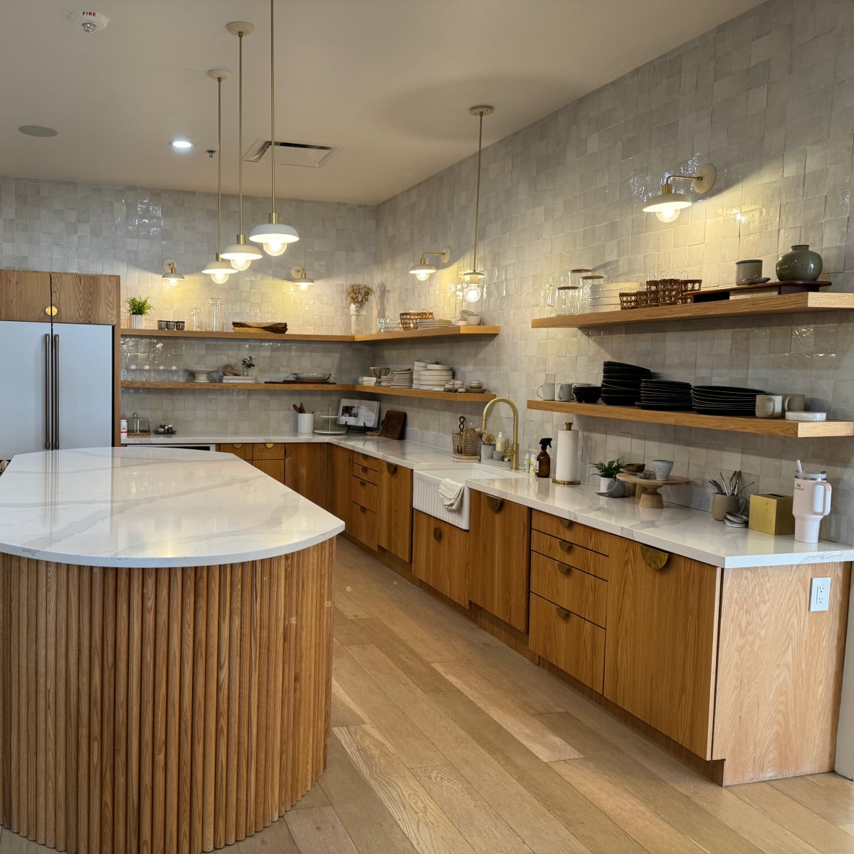 A modern kitchen features a large island with a white countertop, wooden cabinetry, open shelving with dishware, and multiple light fixtures.